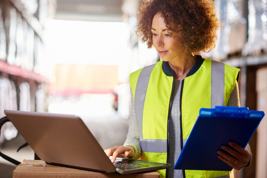 Female supervisor in high-visibility vest reviewing safety and impairment training notes on a laptop in a warehouse.